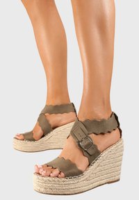 Sandales à talon en toile vert olive avec des brides à bords festonnés, un détail de boucle et une semelle en jute texturée. Design à bout fermé, avec une finition lisse.