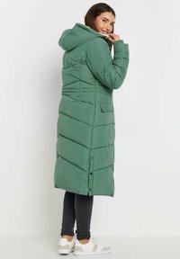 Lång grön puffjacka med huva, med chevron-sömmar, tryckknappar och en sidoficka. Bärs över mörka byxor och vita sneakers.