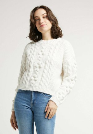 Pull en tricot blanc avec un motif en torsades et en boules, doté d'un col rond et d'une longueur crop, associé à un jean en denim bleu.