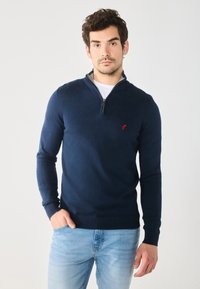Tmavě modrý pletený svetr na zip s červeným logem, s dlouhými rukávy, vysokým límcem a žebrovanými manžetami a spodním lemováním.