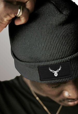 Black knit beanie with ribbed texture, featuring a black patch with a white deer skull logo. The beanie is being adjusted by a hand.