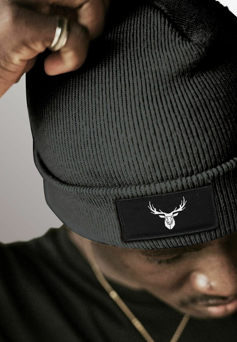 Black knit beanie with ribbed texture, featuring a black patch with a white deer skull logo. The beanie is being adjusted by a hand.