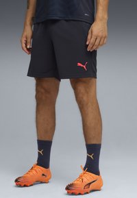 Pantalones cortos deportivos de color azul marino oscuro para hombre con un logo rosa de Puma, combinados con botas de fútbol de color naranja brillante y calcetines azul marino con un logo dorado.