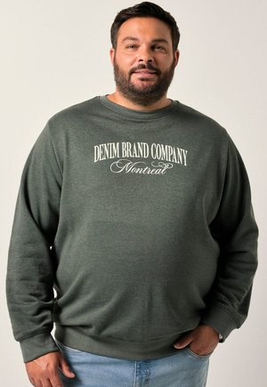 Sudadera de cuello redondo verde con "DENIM BRAND COMPANY Montreal" impreso en blanco. Textura suave, ajuste relajado, combinada con jeans de color azul claro.