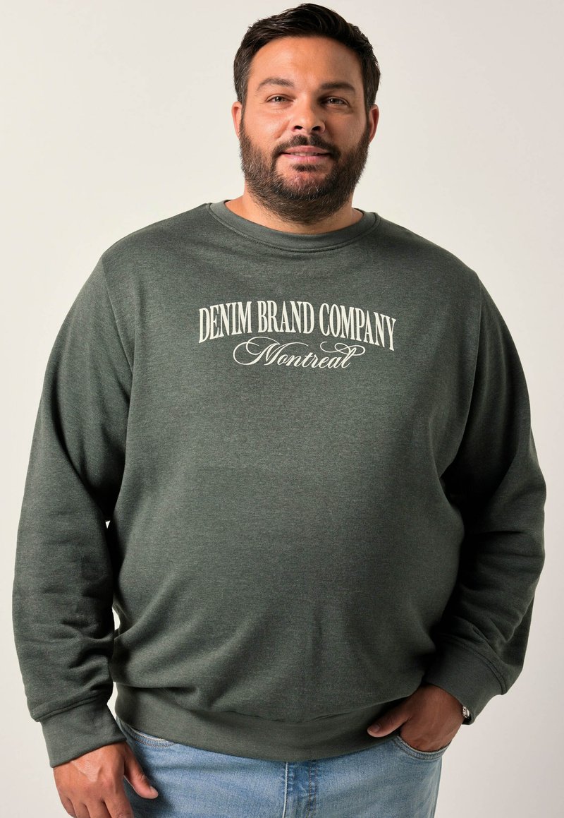 Felpa verde con collo rotondo con la scritta "DENIM BRAND COMPANY Montreal" stampata in bianco. Tessuto morbido, vestibilità rilassata, abbinata a jeans azzurri.