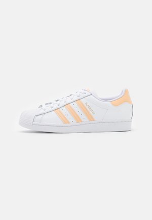 Witte Adidas Superstar sneaker met licht perzikkleurige drie strepen, hielkap, veters en shell toe ontwerp op een effen witte achtergrond.