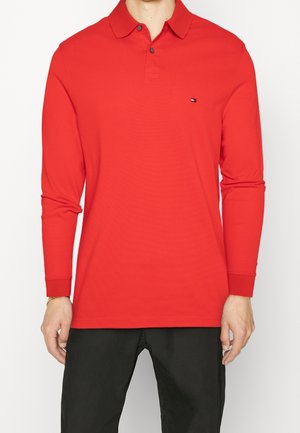Mann trägt ein rotes Langarm-Poloshirt und schwarze Hose, steht mit entspannten Armen an den Seiten, kleines Logo auf der Brust.