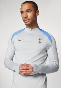 Graues langärmliges Sportoberteil mit blauen Schulterakzenten, Reißverschlusskragen und schwarzem Nike-Logo. Verfügt über Daumenlöcher an den Manschetten für zusätzlichen Komfort.