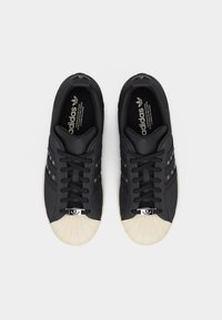 Ζευγάρι μαύρα αθλητικά παπούτσια Adidas με λευκά καουτσούκ ανοιχτά δάχτυλα, μαύρα κορδόνια και το λογότυπο Adidas στη σόλα, από πάνω σε λευκό φόντο.