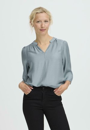 Hellblaue, langärmlige Bluse mit V-Ausschnitt und gerafften Schultern, aus glattem Stoff gefertigt; kombiniert mit schwarzen, figurbetonten Hosen.