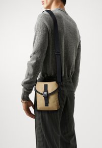Beige canvas crossbody bag with black adjustable strap, snap closure, and accent stitching. Textured, compact, suitable for casual use.