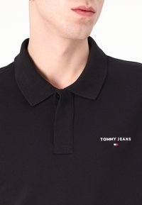 Polo de culoare neagră, fabricat din bumbac, cu guler clasic și placket cu două butoane. Prezintă logo-ul brodat în alb "TOMMY JEANS" și un mic emblem colorat.