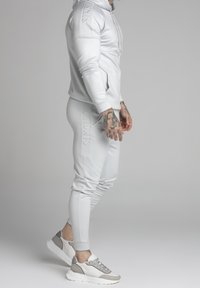 Tuta atletica grigio chiaro con felpa aderente e pantaloni sportivi, caratterizzata da logo impresso, texture liscia e abbinata a sneakers grigie e bianche.