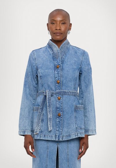 Jachetă din denim de culoare bleu deschis, cu guler mandarin, închidere cu nasturi, talie cu curea și două buzunare frontale. Are cusături și textură subtile.
