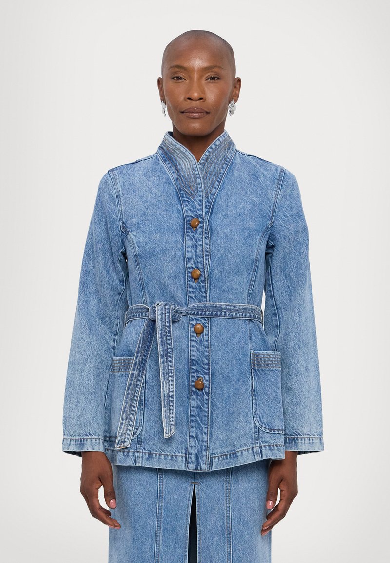 Lys blå denimjakke med ståkrage, knappeåpning, belte i midjen og to frontlommer. Har subtil søm og tekstur.