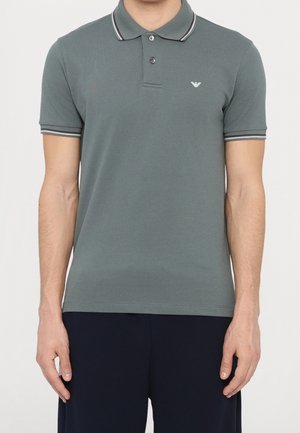 Homme portant un polo à manches courtes gris-vert avec col et poignets rayés blancs et noirs, et pantalon foncé.