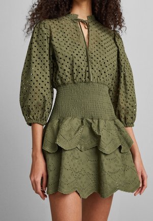 Robe vert olive avec broderie à œillets, taille froncée, jupe à volants et étages, manches bouffantes trois-quarts, et encolure à nouer.