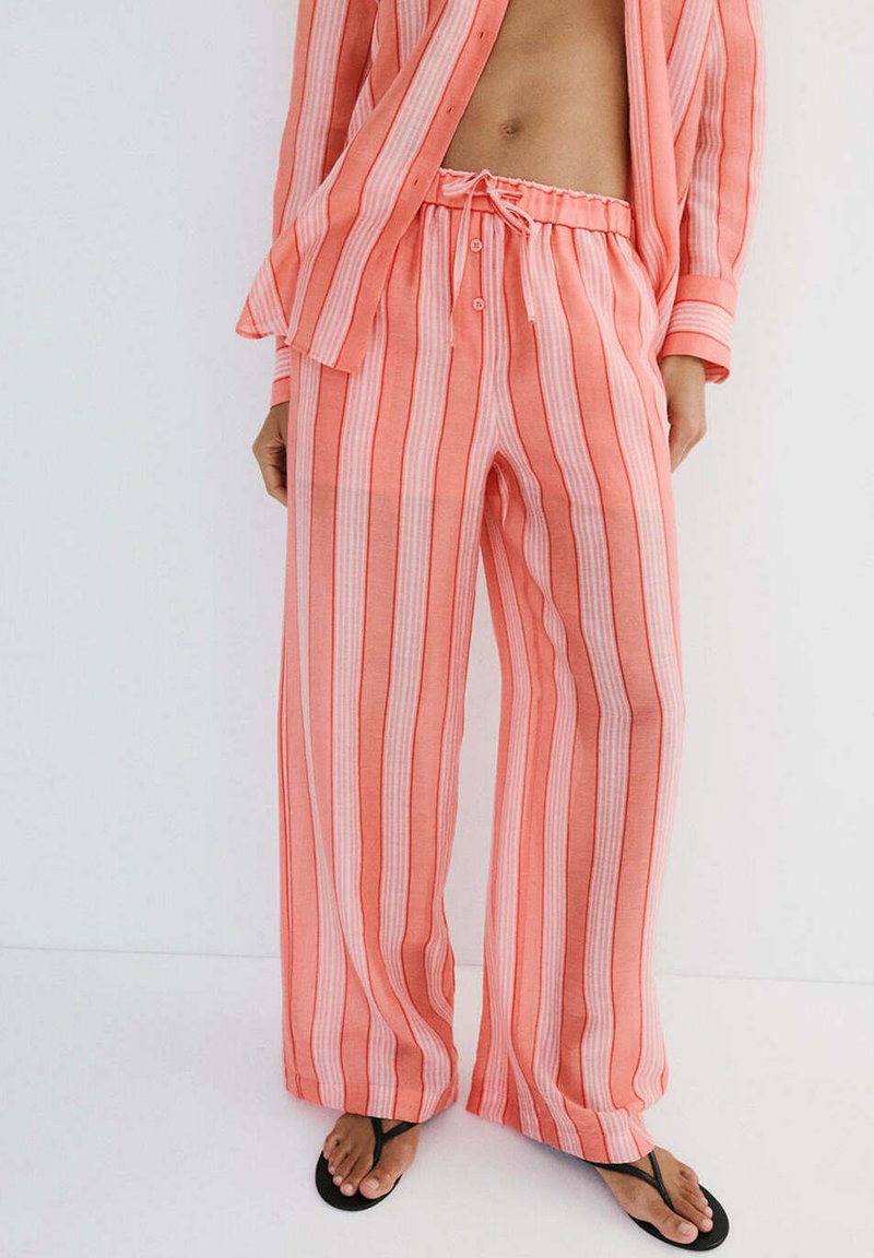 Personne portant un pantalon ample rayé verticalement corail et blanc et une chemise ouverte assortie, associée à des tongs noires, debout sur fond blanc.