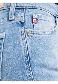 Hele sinised teksapüksid lähivaates, näidates esist taskut punase ja valge tähelogoga sildiga ning metallist tihvti detailiga.