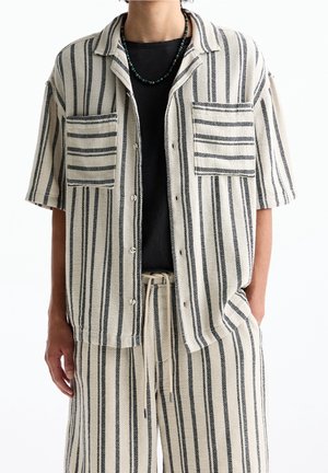 Person trägt ein beiges Hemd mit kurzen Ärmeln und Knöpfen sowie passende Shorts mit vertikalen schwarzen Streifen, ein schwarzes Unterhemd und eine Perlenkette.