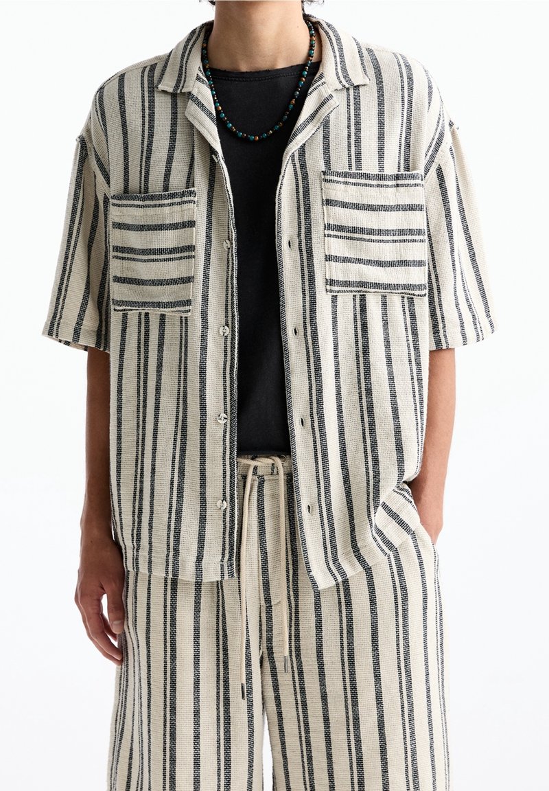 Personne portant une chemise beige à manches courtes avec des boutons et un short assorti à rayures noires verticales, un sous-shirt noir et un collier de perles.