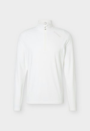 Hombre de pie de frente, vistiendo una camisa blanca de manga larga, pantalones negros con cinturón y zapatillas blancas sobre un fondo liso.