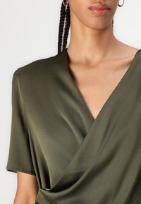 Olīvu zaļas krāsas apģērbs ar īsām piedurknēm, izgatavots no gludas, vieglas auduma, ar dziļu V veida kakla izgriezumu un drapētu priekšpusi.