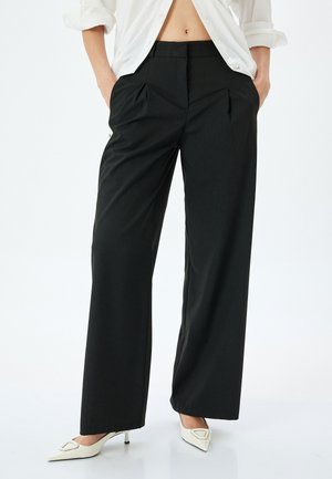 Zwarte wijde broek met plooien in de taille, gemaakt van textuurmateriaal. Gecombineerd met puntige witte hakken en een losse witte blouse.