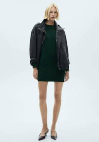 Giacca di pelle nera, oversize, sovrapposta a un vestito a maglia verde scuro. La modella indossa tacchi alti bordeaux con punta affilata. Texture lisce evidenti.