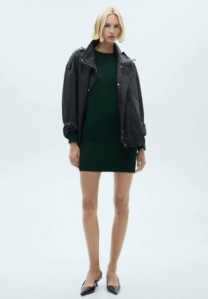 Veste en cuir noire, surdimensionnée, superposée à une robe pull vert foncé. Le mannequin porte des escarpins bordeaux à bout pointu. Textures lisses évidentes.