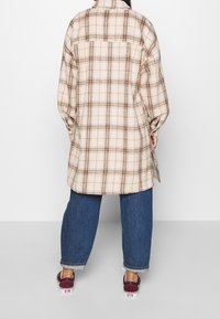 Persona in piedi di spalle, indossa una camicia lunga a quadri beige e marrone, jeans blu e sneakers bordeaux.