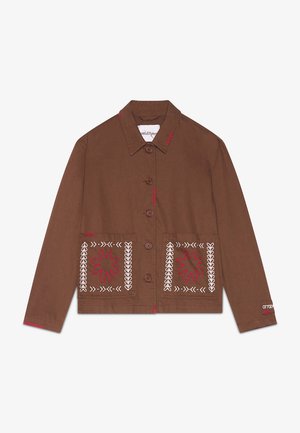 Vrouw draagt een bruine jas met knopen en rood-witte borduursels met bijpassende bruine shorts, staand tegen een effen achtergrond.