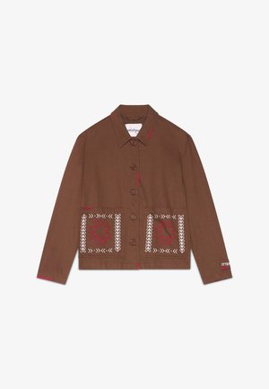 Vrouw draagt een bruine jas met knopen en rood-witte borduursels met bijpassende bruine shorts, staand tegen een effen achtergrond.