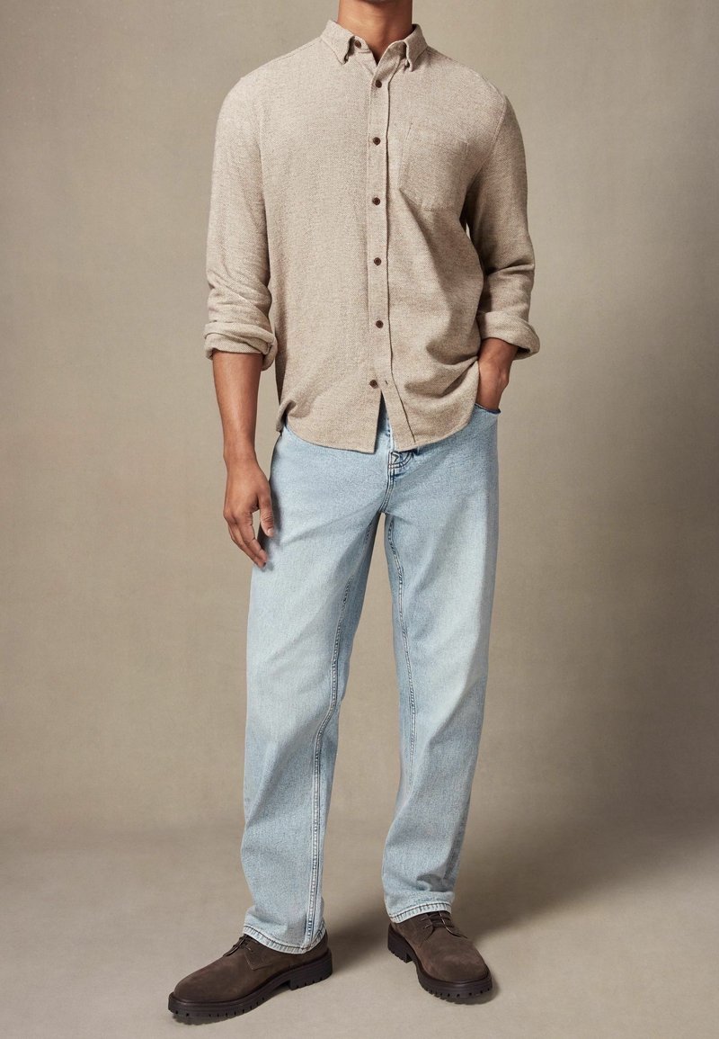 Homme portant une chemise beige à manches longues boutonnée, un jean bleu clair et des chaussures marron foncé à lacets, debout avec une main dans la poche.