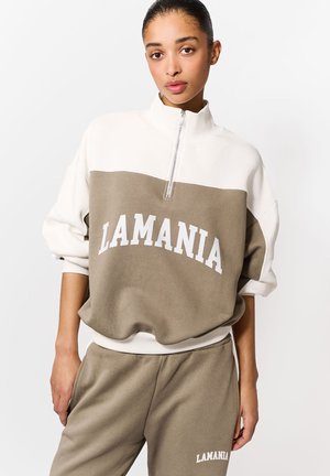 Světle krémový a khaki pulovr s čtvrtinovým zipem, s nápisem "LAMANIA" bílými písmeny přes hruď. Měkká, strukturovaná tkanina.