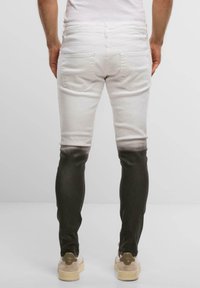 Muž v slim-fit džínách s bílým vrškem a černými dolními nohavicemi, stojící zády k nám na jednobarevném světlém pozadí.