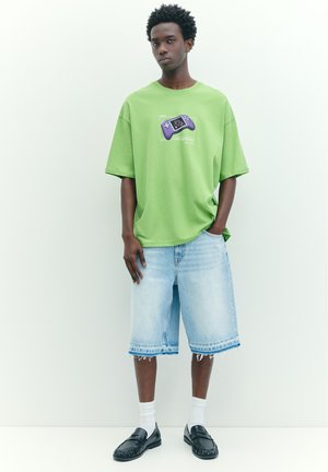Junger Mann steht, trägt ein grünes T-Shirt mit einem violetten Gamecontroller-Motiv, helle Denim-Langshorts, weiße Socken und schwarze Loafers.