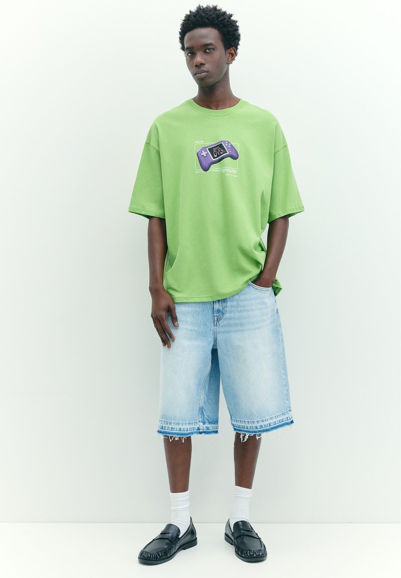 Jeune homme debout portant un t-shirt vert avec un motif de manette de jeu violet, un short long en jean clair, des chaussettes blanches et des mocassins noirs.