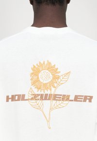 Valkoinen puuvillapaita, jossa on keltainen brodeerattu auringonkukkakuvaus, joka on yksityiskohtainen, sekä sana "HOLZWIELER" ruskealla.