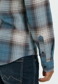 Blå og hvit rutete skjorte med brune detaljer; har lange ermer, knappede mansjetter og en buet hem. Brukt med mørk denimjeans.
