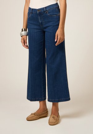 Weite, dunkelblaue Jeans mit hoher Taille, ausgestattet mit Fronttaschen, einer Knopfleiste und kontrastierenden Nähten. Kombiniert mit beigen Ballerinas.