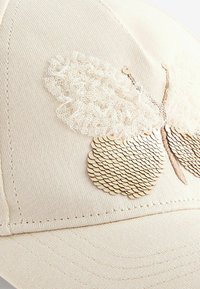Gorra beige con un diseño de mariposa texturizado, con tela tipo encaje y lentejuelas metálicas, resaltando detalles intrincados y forma.