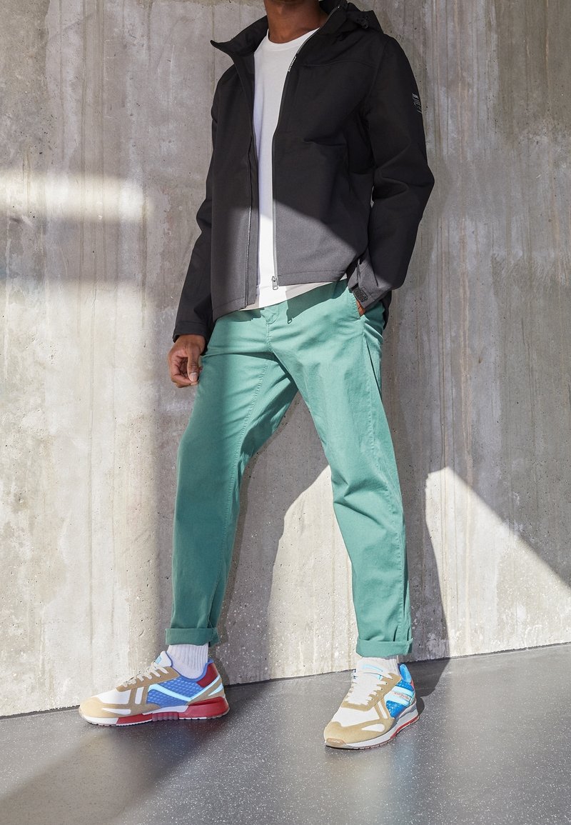 Černá zipová bunda, bílá tričko, mátově zelené kalhoty a vícebarevné tenisky s modrými, červenými a hnědými akcenty, na pozadí betonové zdi.