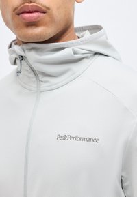 Κοντινό πλάνο ενός ατόμου που φοράει ζακέτα με φερμουάρ Peak Performance σε ανοιχτό γκρι χρώμα, με κορδόνια και εμφανές λογότυπο στο στήθος.