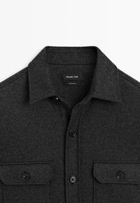 Ciemnoszara koszula wełniana z zapięciem na guziki, z wyraźnym kołnierzykiem i dwiema przednimi kieszeniami na klapki, oznaczona etykietą "Massimo Dutti" i wyprodukowana w Turcji.