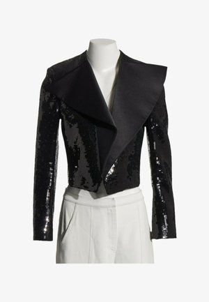 Chaqueta corta negra con lentejuelas y amplias solapas de satén mostrada en un maniquí blanco que viste pantalones blancos.