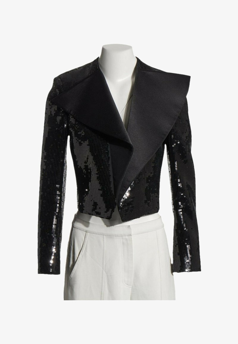 Veste courte noire à sequins avec larges revers en satin présentée sur un mannequin blanc portant un pantalon blanc.
