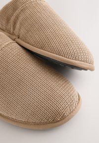 Beige Stoffhausschuhe mit strukturierter Oberfläche, abgerundetem Zehenbereich und genähten Akzenten. Gummisohle sorgt für Griffigkeit und Langlebigkeit.