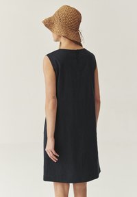 Femme portant une robe noire sans manches et un chapeau de soleil en osier marron, vue de dos contre un fond clair.