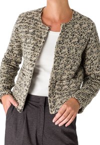 Personne portant une veste texturée beige et noire à motifs sur un haut blanc et un pantalon gris foncé, les mains reposant près des poches.